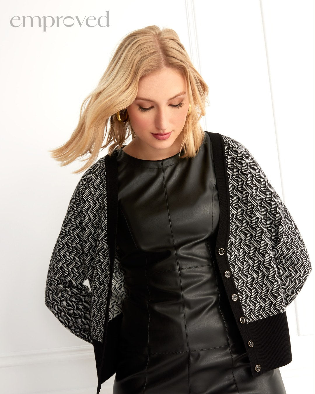 Woman wearing a black leather dress and a patterned cardigan, looking down against a white background