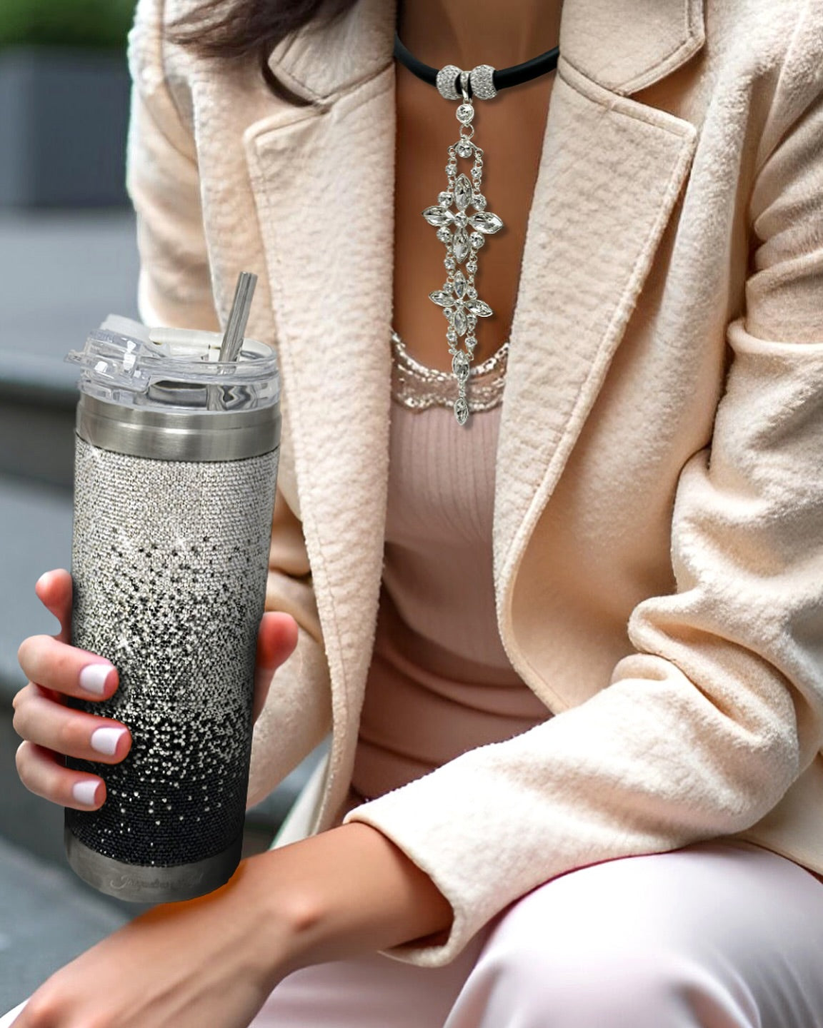 Close-up of a woman wearing a blush-colored outfit with a textured coat, accessorized with a sparkling statement necklace, holding a rhinestone-covered tumbler with a straw.