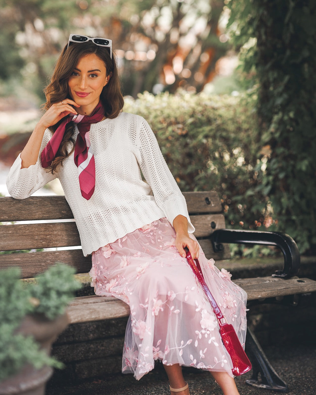 Woman sitting on a park bench wearing a white knit sweater, pink floral embroidered skirt, and a burgundy scarf, holding a red handbag with sunglasses perched on her head.