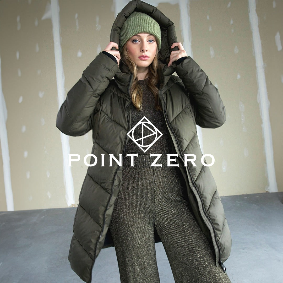 Woman wearing an olive green puffer coat with a matching knit beanie, adjusting her hood while posing against an industrial-style background. 'Point Zero' logo displayed over the image.