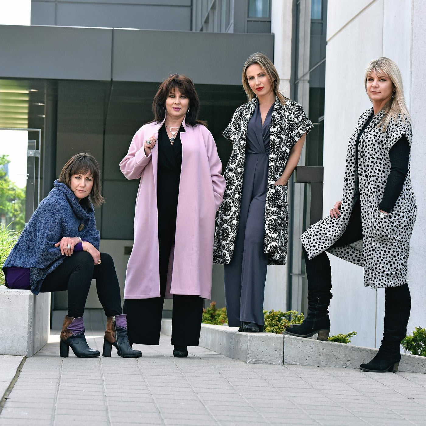Four women posing outdoors in stylish fall and winter outerwear, including coats, vests, and ponchos, in a modern urban setting.