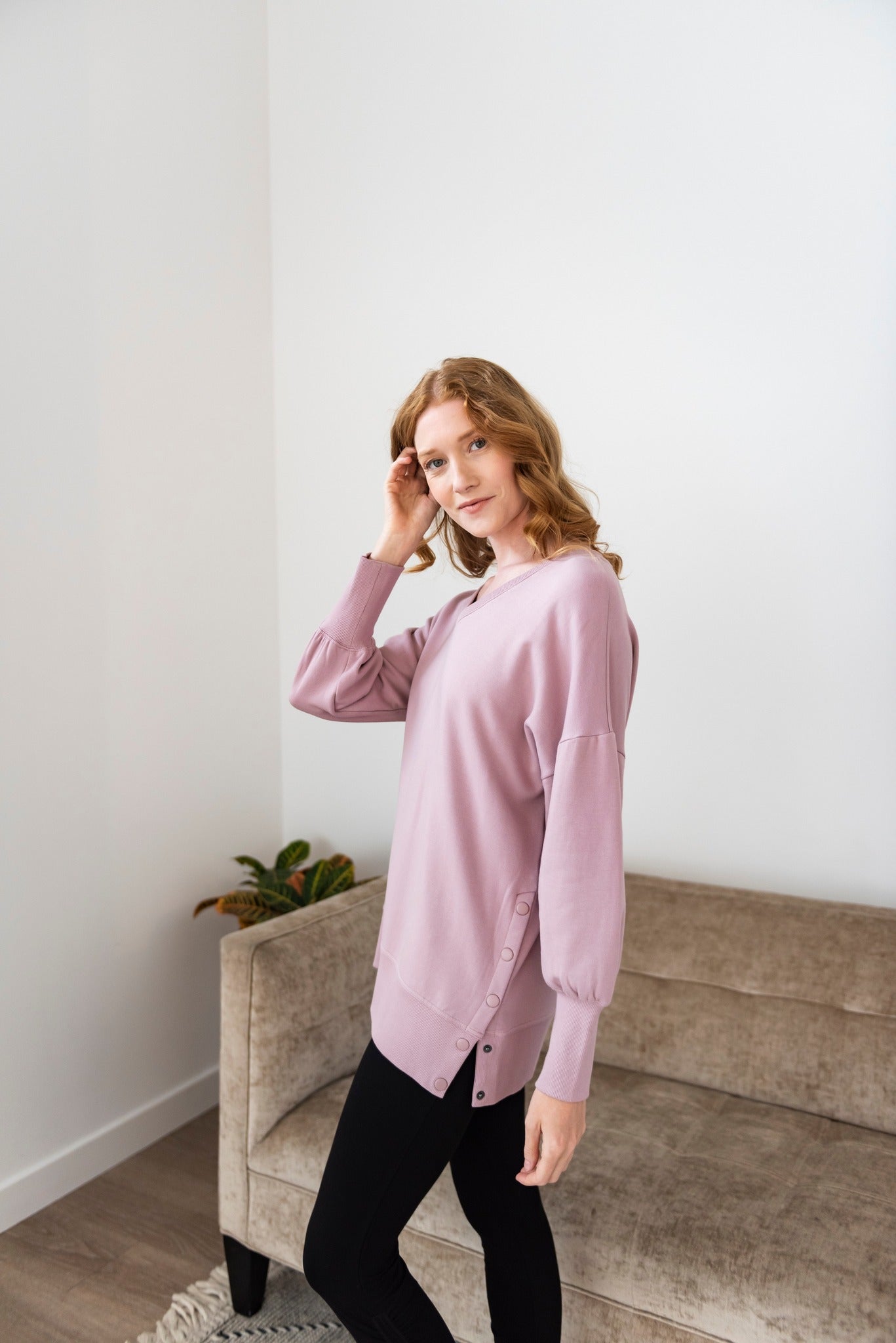 Woman wearing a mauve long-sleeve tunic with button details on the side, paired with black leggings, posing in a modern living space.